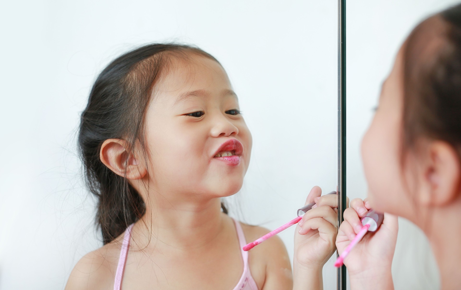 Cute little Asian child girl trying lip gloss near a mirror.